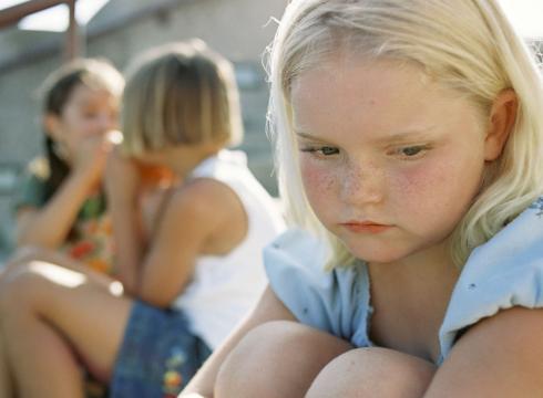 Girl depressed on a playground with girls talking about her.